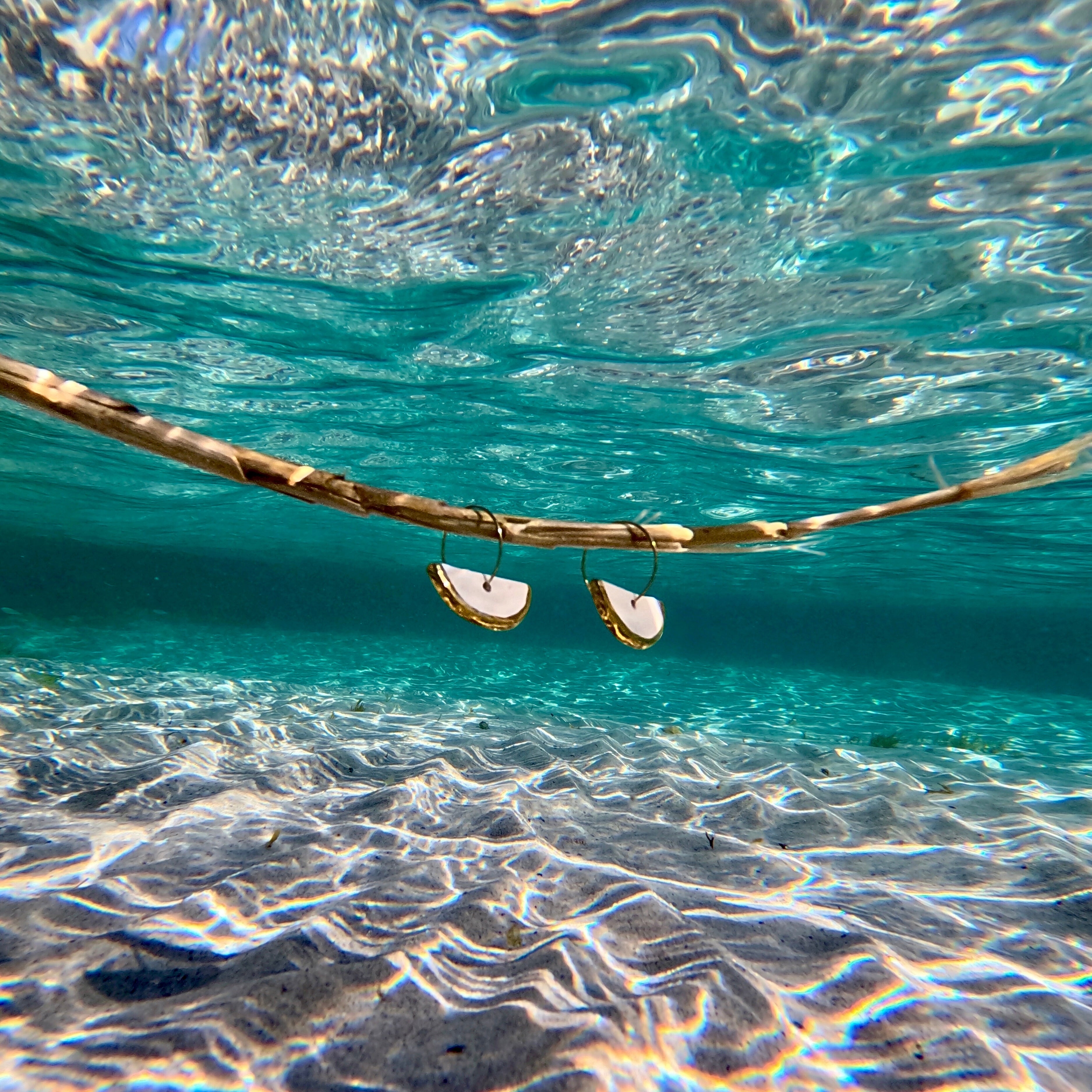 Gold hoop Doraluna earrings by Banceramiche underwater with clear blue water and sand.